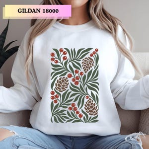 May include: A white crewneck sweatshirt featuring a central graphic print of dark green leaves, brown pinecones, and clusters of red berries. The text GILDAN 18000 is printed in black in the upper left corner of the image.