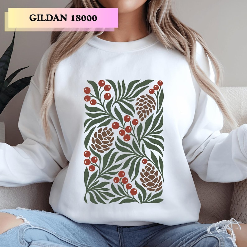 May include: A white crewneck sweatshirt featuring a central graphic print of dark green leaves, brown pinecones, and clusters of red berries. The text GILDAN 18000 is printed in black in the upper left corner of the image.