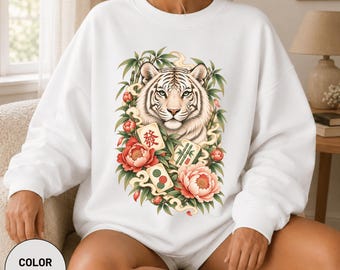 Könnte beinhalten: Ein weißes Sweatshirt mit Rundhalsausschnitt und einem zentralen Aufdruck eines weißen Tigers, umgeben von grünen Bambusstangen, rosa Pfingstrosen und drei Mahjong-Steinen. Die Steine zeigen rote und grüne Symbole sowie das chinesische Zeichen fa.