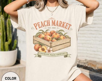 May include: An ivory short-sleeve t-shirt featuring a graphic of a wooden crate filled with peaches. The text reads PEACH MARKET EST. 1986 and PREMIUM PEACH GROWERS. The design includes orange and green accents, showcasing a collection of fresh fruit.