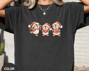 Könnte beinhalten: Ein anthrazitfarbenes Kurzarm-T-Shirt mit einer Grafik von drei Cartoon-Pinguinen, die rote Weihnachtsmützen tragen. Der linke Pinguin hält eine Zuckerstange, der mittlere ein rotes Herz und der rechte Pinguin ein rotes Geschenkpaket.