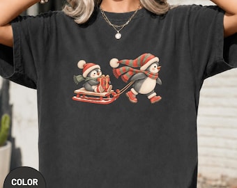 Könnte beinhalten: Ein anthrazitfarbenes T-Shirt mit einer Grafik von zwei Pinguinen. Ein Pinguin trägt eine rot-grüne Mütze und einen Schal und zieht einen Holzschlitten. Der andere Pinguin sitzt auf dem Schlitten, trägt eine rote Mütze und Schal und hält ein Geschenk.