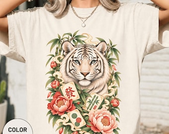 Könnte beinhalten: Ein elfenbeinfarbenes T-Shirt mit einem Grafikdruck eines weißen Tigerkopfes, umgeben von grünen Bambusstangen, roten und rosafarbenen Pfingstrosen sowie drei Mahjong-Steinen. Der mittlere Stein zeigt ein rotes chinesisches Schriftzeichen.