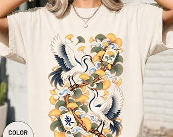 Könnte beinhalten: Ein elfenbeinfarbenes T-Shirt mit einem Grafikdruck von zwei weißen Kranichen mit blau-schwarzen Flügeldetails, die auf Ästen sitzen. Das Design zeigt gelbe Ginkgoblätter, blaue Wirbelwolken und zwei Mahjong-Steine, einer davon mit dem blauen Schriftzeichen 東.