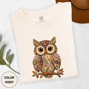 May include: An ivory t-shirt featuring a graphic of an owl perched on a branch. The owl is decorated with various patterns including leaves, mushrooms, moons, and plaid in shades of brown, purple, green, and gold. The shirt has a crew neckline.
