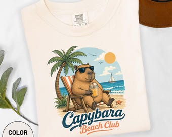 Könnte beinhalten: Ein elfenbeinfarbenes T-Shirt mit einer Grafik eines Capybaras mit Sonnenbrille, das in einem Strandstuhl sitzt und ein Getränk hält, dazu eine Palme, das Meer, ein Segelboot und die Sonne. Der Text lautet Capybara Beach Club.