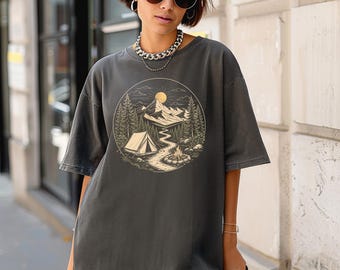 Könnte beinhalten: Ein anthrazitfarbenes Kurzarm-T-Shirt mit einem kreisförmigen Grafikdruck auf der Brust. Das Design zeigt eine Berglandschaft mit einem Zelt, einem gewundenen Pfad, einem Lagerfeuer, Tannen und einer Sonne, alles in hellbeiger Linienführung.