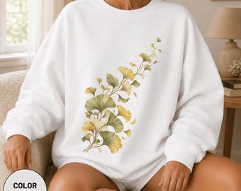 May include: A white crewneck sweatshirt featuring a botanical illustration of ginkgo biloba leaves in shades of yellow and green. The design extends diagonally across the front of the garment, showcasing fan-shaped leaves on thin stems.