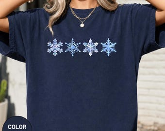 Könnte beinhalten: Ein marineblaues T-Shirt mit kurzen Ärmeln, das in der Mitte der Brust eine horizontale Reihe aus vier verschiedenen, filigranen Schneeflockenmustern in hellblauen, weißen und zartvioletten Farbtönen zeigt.