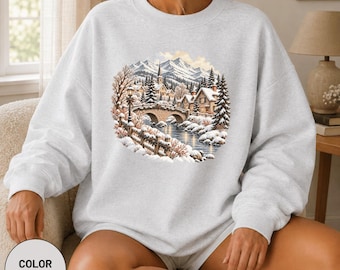 Könnte beinhalten: Ein aschgraues Sweatshirt mit Rundhalsausschnitt und einem kreisförmigen Grafikdruck einer verschneiten Winterlandschaft. Die Abbildung zeigt eine Steinbrücke über einen Fluss, Tannenbäume, Häuser und Berge im Hintergrund.