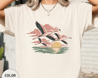 Könnte beinhalten: Ein elfenbeinfarbenes T-Shirt mit einem Grafikdruck von drei rosa Flamingos, die bei Sonnenuntergang über ein ruhiges Meer fliegen. Das Design zeigt blassrosa Wolken, eine gelbe Sonne und grünes Wasser und bildet eine ruhige Küstenszene.