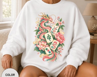Könnte beinhalten: Ein weißes Sweatshirt mit Rundhalsausschnitt und einer zentralen Grafikillustration eines gewundenen Drachen, umgeben von rosa Lotusblüten, grünen Blättern und vier Mahjong-Steinen. Die Steine zeigen verschiedene Symbole, darunter ein grünes Schriftzeichen.