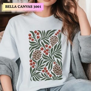 May include: A white t-shirt featuring a graphic print of dark green leaves, brown pinecones, and red berries. The text BELLA CANVAS 3001 is printed in the upper left corner of the image. The shirt has a crew neckline and short sleeves.