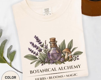 Könnte beinhalten: Ein elfenbeinfarbenes T-Shirt mit einer Grafik einer violetten Zaubertrankflasche, umgeben von Lavendelzweigen, grünen Blättern und zwei Pilzen. Unter der Illustration steht der Text BOTANICAL ALCHEMY sowie HERBS • BLOOMS • MAGIC.