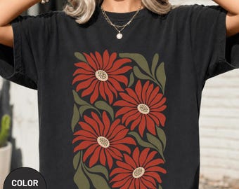 May include: A black short-sleeved t-shirt featuring a graphic print of four large, rust-colored flowers with cream-colored centers and dark olive green leaves. The design is arranged in a rectangular block pattern on the front of the garment.