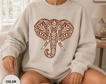 Könnte beinhalten: Ein sandfarbenes Sweatshirt mit Rundhalsausschnitt und einer großen, detaillierten braunen Grafik eines Elefantenkopfes. Das Elefantenmotiv ist mit komplexen Blatt- und geometrischen Mustern gefüllt und mittig auf der Brust platziert.
