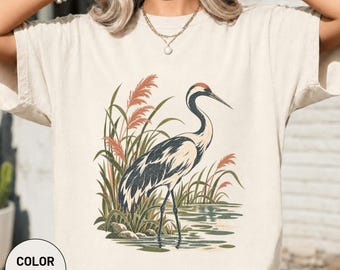 Könnte beinhalten: Ein elfenbeinfarbenes T-Shirt mit einer grafischen Illustration eines Kranichs, der im Wasser zwischen hohen Schilfhalmen steht. Der Vogel ist in Schwarz-, Weiß- und Grautönen gehalten, während das Schilf in gedeckten Orange- und Grüntönen erscheint.