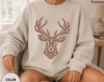 Könnte beinhalten: Ein sandfarbenes Sweatshirt mit Rundhalsausschnitt und einer großen, braunen, detaillierten Grafik eines Hirschkopfes auf der Brust. Das Hirschdesign besteht aus verschiedenen geometrischen Mustern, darunter Blätter, Punkte und Linien.