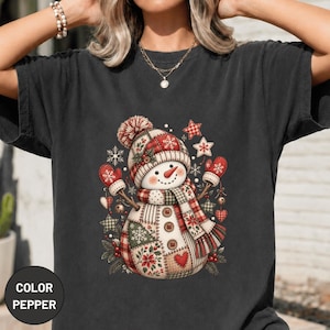 May include: A charcoal gray t-shirt featuring a central graphic of a snowman wearing a red and white plaid hat and scarf. The snowman has a patchwork body design with snowflake and floral patterns, red mittens, and three brown buttons.
