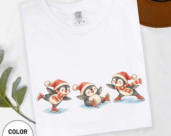 Könnte beinhalten: Ein weißes T-Shirt mit einer Grafik von drei Cartoon-Pinguinen, die rote Wintermützen und gestreifte Schals tragen. Die Pinguine sind beim Eislaufen auf einer hellblauen Fläche dargestellt. Das T-Shirt hat ein Comfort Colors Etikett am Kragen.