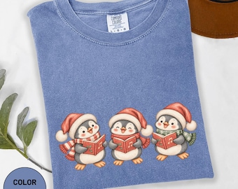 Könnte beinhalten: Ein jeansblaues T-Shirt mit Rundhalsausschnitt und einem Comfort Colors Markenetikett. Die Grafik zeigt drei Cartoon-Pinguine mit roten Weihnachtsmützen und Schals, die jeweils ein rotes Liederbuch mit aufgedruckten Musiknoten in den Händen halten.