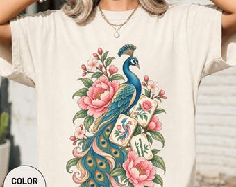 Könnte beinhalten: Ein elfenbeinfarbenes T-Shirt mit einem Grafikdruck eines blauen Pfaus, umgeben von rosa Pfingstrosen und grünen Blättern. Das Design enthält drei rechteckige Kacheln, eine mit einem Pfau, eine mit einer rosa Blume und eine mit grünen Bambusstangen.