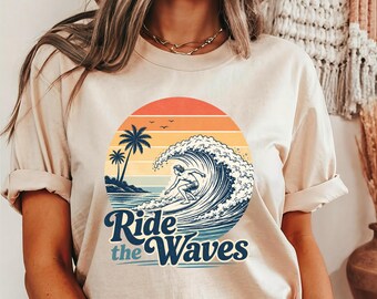 Könnte beinhalten: Ein beiges T-Shirt mit kurzen Ärmeln und einem kreisförmigen Aufdruck, der einen Surfer auf einer großen Welle zeigt. Das Design enthält Palmen, einen Sonnenuntergang und den Text Ride the Waves in dunkelblauer Schrift. Das Shirt hat einen Rundhalsausschnitt.