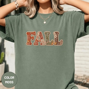 May include: A moss green short-sleeve t-shirt featuring the word FALL printed across the chest. The letters are filled with a multi-colored paisley and floral pattern in shades of orange, brown, and green, outlined with a white stitched border.
