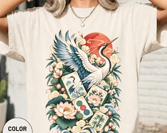 Könnte beinhalten: Ein elfenbeinfarbenes T-Shirt mit einem Grafikdruck eines Kranichs mit blau-weißen Flügeln, umgeben von Lotusblumen, grünen Blättern, wirbelnden Wolken, einer roten Sonne und mehreren Mahjong-Steinen mit Kranich- und Bambusmotiven.