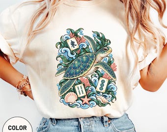 Könnte beinhalten: Ein elfenbeinfarbenes T-Shirt mit einem Grafikdruck einer Meeresschildkröte, die zwischen grünem Seetang, blauen Wellen, rosa Korallen, Muscheln und drei Mahjong-Steinen schwimmt. Der mittlere Stein zeigt das chinesische Schriftzeichen für Ost.