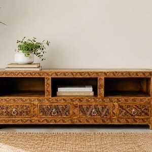 Puede incluir: Un mueble de televisión rectangular de madera con acabado marrón natural, que cuenta con tres compartimentos de almacenamiento abiertos en la parte superior y cuatro cajones debajo. Cada cajón tiene tallas florales y tiradores de anillo de metal.