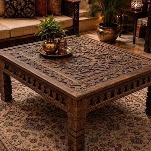 May include: A square wooden coffee table featuring intricate hand-carved floral patterns on the tabletop surface and along the sides. The table has four sturdy legs with carved details and a dark brown finish, supporting a flat, ornate top.