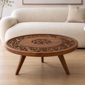 May include: A round, low-profile coffee table crafted from medium-brown wood. The tabletop features an intricate, hand-carved floral relief pattern encircling a central medallion. The table is supported by three tapered, angled wooden legs.