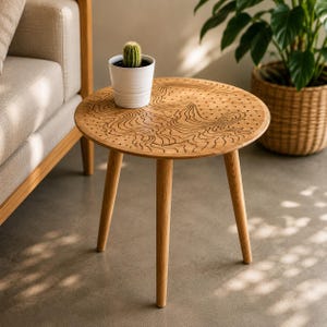 Pode incluir: Uma mesa lateral redonda de madeira com três pernas cónicas e acabamento castanho claro. O tampo apresenta um padrão gravado escuro de mapa topográfico com linhas onduladas e pequenos pontos. Um pequeno cacto verde está num vaso branco.