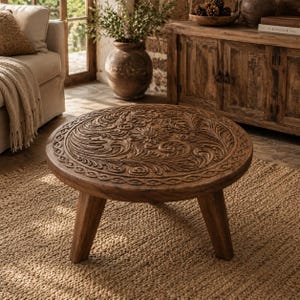 Peut inclure: Une table basse ronde en bois présentant des motifs floraux et des volutes finement sculptés à la main sur le plateau. La table est soutenue par trois pieds fuselés robustes et possède une finition brune naturelle avec un grain de bois visible.