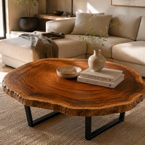 Op de afbeelding: Een ronde salontafel met een natuurlijk boomstam-houten blad met een rijke bruine afwerking en zichtbare nerven. De tafel wordt ondersteund door twee zwarte, U-vormige metalen poten. Een kleine keramische kom en twee boeken liggen op het blad.