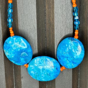 May include: A necklace featuring three large, oval-shaped blue stones with marbled patterns. The stones are separated by small, bright orange beads and translucent blue beads. The necklace strand consists of alternating orange and blue beads.