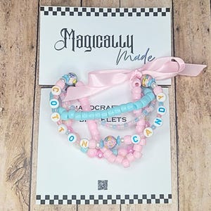 May include: A set of multi-strand beaded bracelets featuring pink and light blue plastic beads, floral patterned accent beads, and letter beads spelling COTTON CANDY. The bracelets are tied with a light pink ribbon on a white display card.