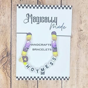 以下が含まれることがあります： Magically Made Handcrafted Braceletsと書かれた白い台紙に固定されたビーズブレスレット。黄色、紫、ダークグレーの平らなディスクビーズ、ハート付きの茶色いアイスキャンディーのチャーム、そしてHOTMESSと綴られた白い文字ビーズで構成されています。