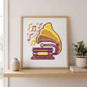 May include: A cross-stitch embroidery pattern in a wooden frame featuring a vintage gramophone in shades of yellow, gold, and purple. Musical notes in yellow and orange float above the gramophone, which is set against a plain white fabric background.