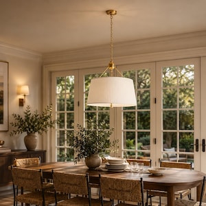 May include: A dining room set featuring a large oval wooden table surrounded by six chairs with black metal frames and woven seats. Above the table hangs a large, white, pleated drum pendant light with a gold-toned chain and hardware.