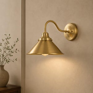 May include: A wall-mounted lamp featuring a brushed brass finish with a conical shade and a curved arm. The fixture includes a circular wall plate and a single downward-facing light source that casts a soft glow on the surface below the lamp.