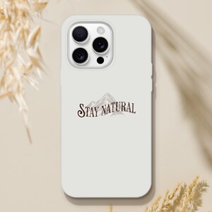 Könnte beinhalten: Eine matte, cremeweiße Smartphone-Hülle mit einer dunkelbraunen Grafik eines Gebirgszuges und dem Text STAY NATURAL in einer stilisierten Serifenschrift, die mittig auf der Rückseite platziert ist und eine Aussparung für drei Kameras aufweist.