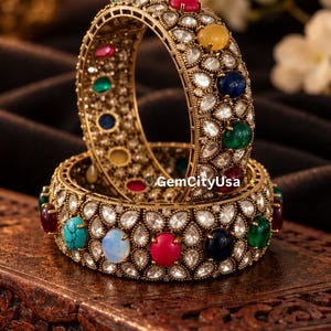 May include: Two gold-toned metal bangles featuring a repeating pattern of teardrop-shaped clear stones and round cabochon stones in red, blue, green, yellow, and turquoise. The text GemCityUsa is visible on the surface of the bangles.