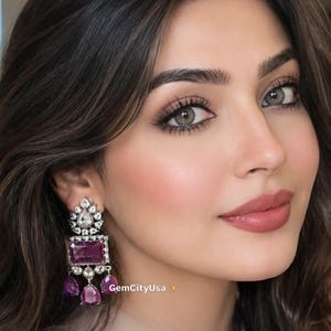 May include: A dangling earring featuring a rectangular purple gemstone at the center, topped with a floral-shaped cluster of clear white stones, and finished with three smaller pear-shaped purple drops. The text GemCityUsa is visible.