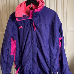 Puede incluir: Una chaqueta Columbia de color morado con detalles en rosa brillante en el cuello, los paneles laterales y los tiradores de las cremalleras. La chaqueta cuenta con cuello alto, cierre frontal de cremallera con solapa y mangas largas con puños elásticos.