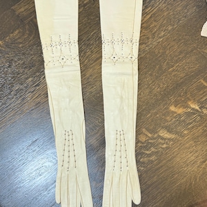 May include: A pair of long, cream-colored opera gloves featuring intricate beadwork patterns. The design includes a scalloped beaded motif near the forearm and vertical beaded lines extending from the wrist toward the fingers on each glove.