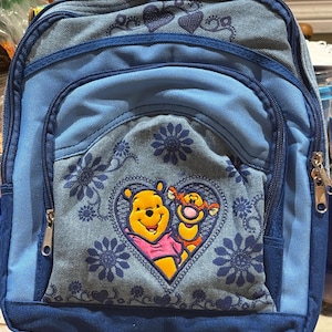 May include: A light and dark blue denim backpack featuring an embroidered heart design on the front pocket. Inside the heart are Winnie the Pooh and Tigger. The backpack is decorated with dark blue floral patterns and has multiple zippered compartments.