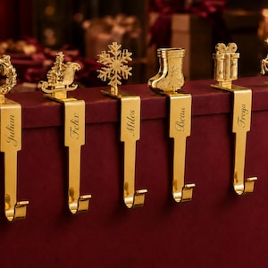 May include: Six gold-toned metal stocking holders featuring festive toppers including a wreath, sleigh, snowflake, stocking, gift, and reindeer. Each vertical arm is engraved with a name: Julian, Felix, Miles, Beau, Freya, and Joy.