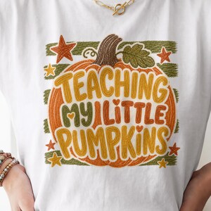 May include: A white short-sleeved t-shirt featuring a graphic of an orange pumpkin with green leaves and stars. The text reads "TEACHING MY LITTLE PUMPKINS" in yellow and green letters. The design has a textured, embroidered-style appearance.